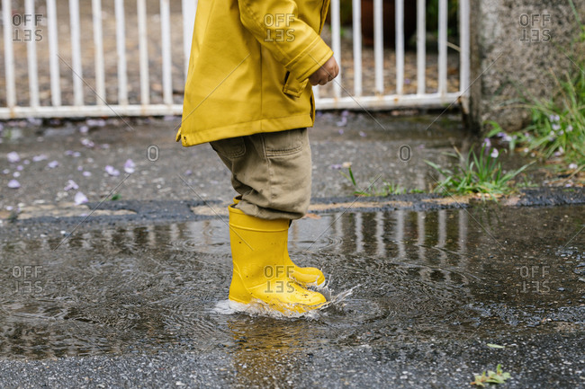 Cropped little playful kid in yellow raincoat and rubber boots walking in puddle and splashing water while having fun after rain