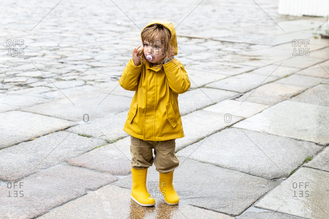 Full body of cute little kid wearing bright yellow raincoat and rubber boots standing on wet pavement and looking away