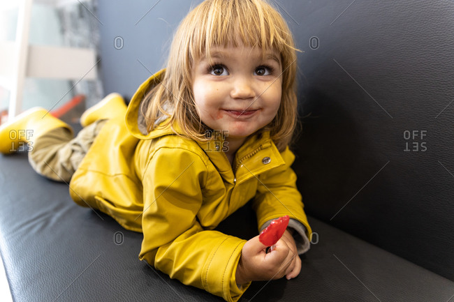 Adorable little kid in yellow raincoat and with dirty mouth lying on sofa in cafe with lollipop and looking away