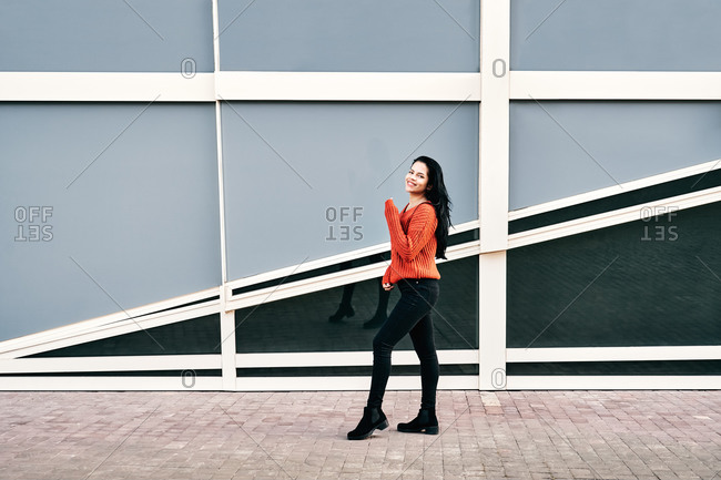 Side view full body of cheerful ethnic female in casual outfit smiling and standing near modern building on windy day in city