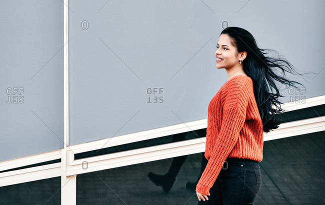 Side view of cheerful ethnic female in casual outfit smiling and walking near modern building on windy day in city