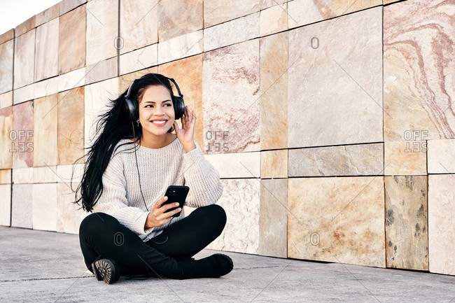 Happy ethnic female smiling and listening to music in headphones while sitting cross legged near marble wall
