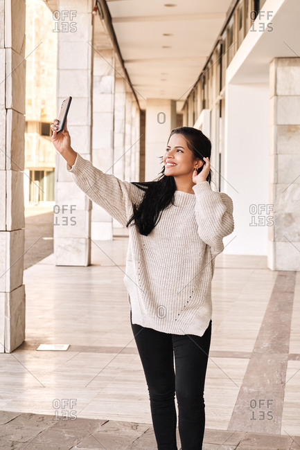 Happy ethnic woman touching hair and taking selfie while standing in marble passage outside building