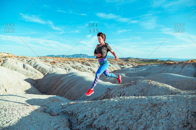 Full body side view of active female athlete in sportswear running and jumping over rocky slope of rough desert hilly terrain under blue sky