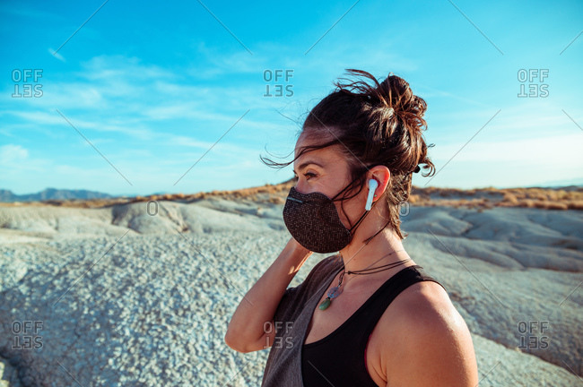 Side view of confident young sportswoman in black protective mask listening to music with wireless earbuds while recreating during training in rough desert badlands looking away