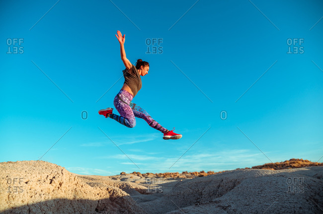 From below full body of active energetic female in sportswear leaping high above sandy slope of desert terrain against blue cloudless sky