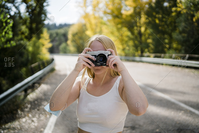 Unrecognizable female photographer covering face with camera while standing on asphalt roadway among forest and taking pictures in sunny autumn day in countryside