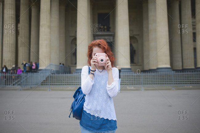 Young red haired female tourist with backpack shooting photo with instant camera while standing against famous Kazan Cathedral in summer day in Saint Petersburg
