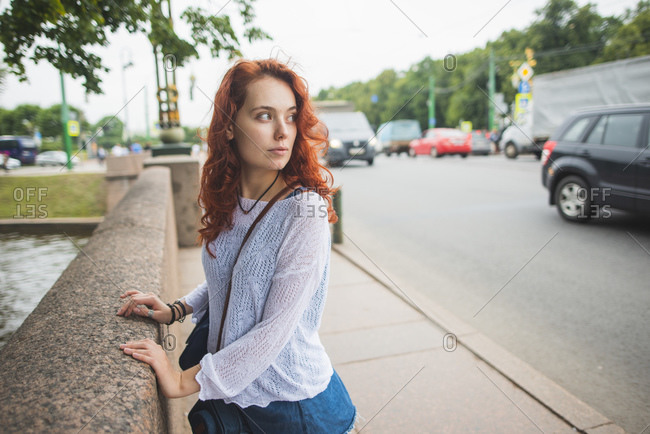 Side view of young red haired female student in casual outfit looking away with interest while standing near stone border on bridge over channel on street of Saint Petersburg