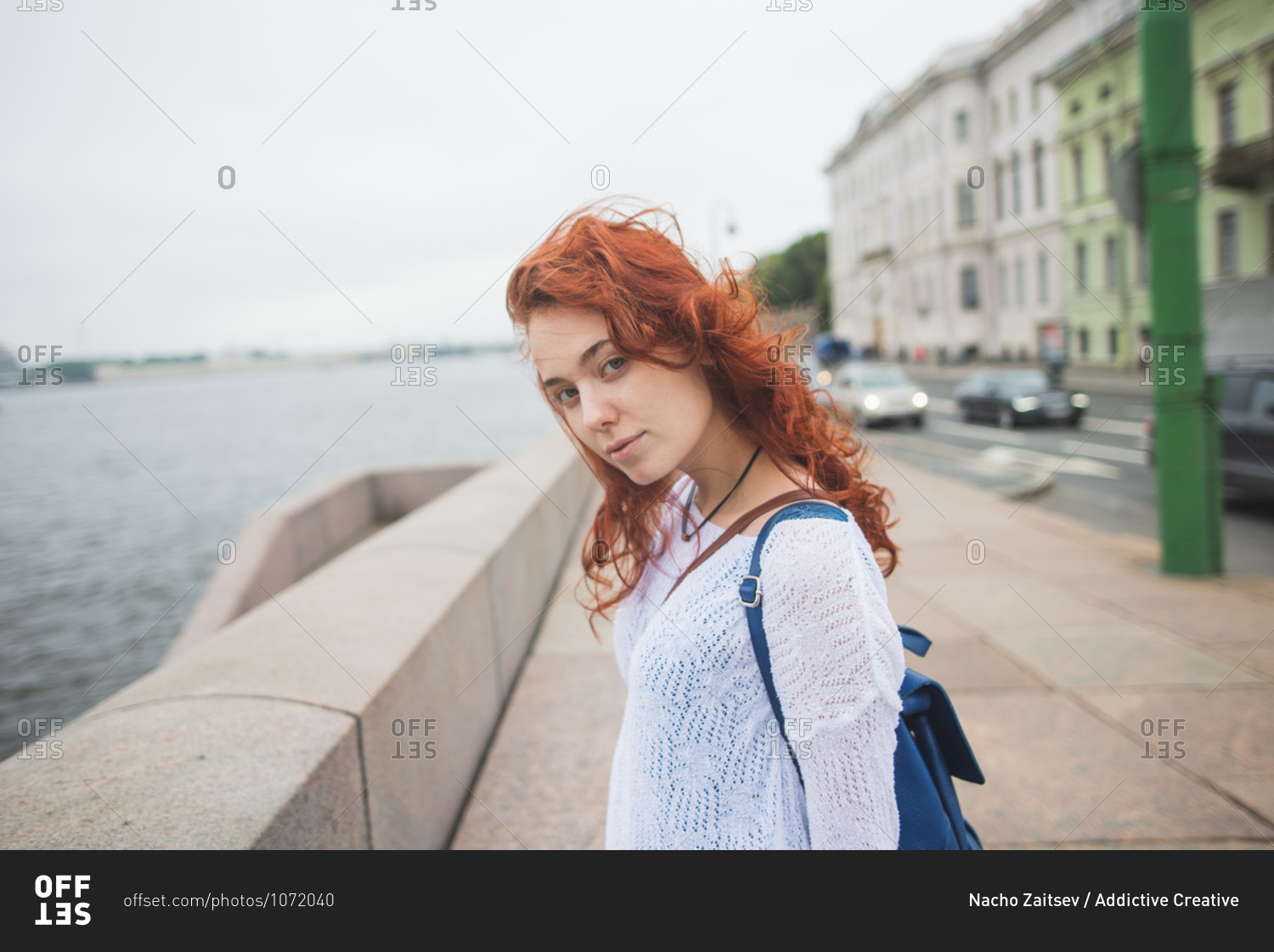 Side view of red haired female tourist with backpack standing near stone fence against river and enjoying wind while resting during sightseeing in cloudy day in Saint Petersburg in Russia