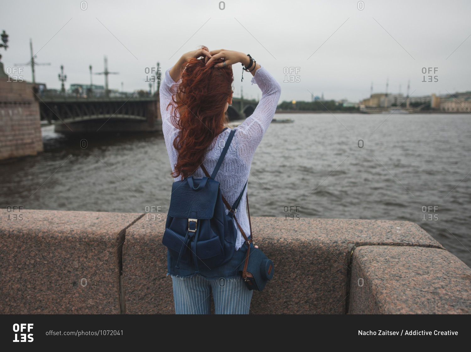 Back view of unrecognizable red haired female tourist with backpack standing near stone fence against river and enjoying wind while resting during sightseeing in cloudy day in Saint Petersburg in Russia