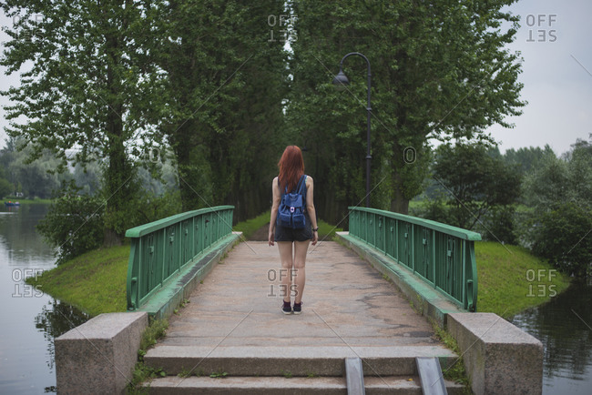 Full body back view of unrecognizable female traveler standing on old footbridge crossing river and leading to small green island while exploring Saint Petersburg neighborhoods in summer day