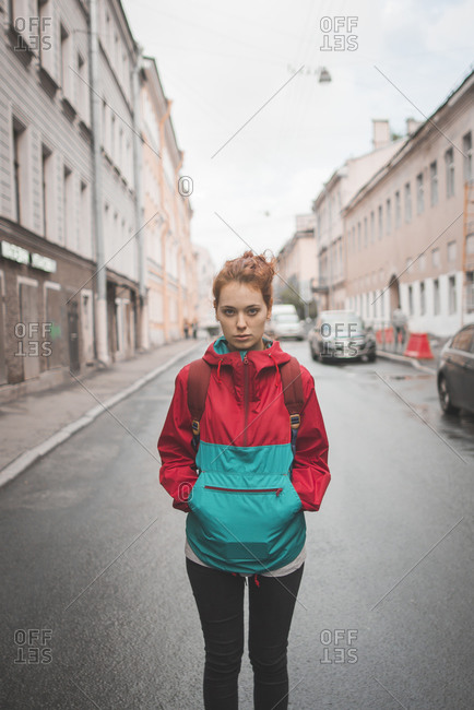 Serious millennial redhead female with curly hair wearing red jacket looking at camera while standing alone against road and historic buildings in windy day in Saint Petersburg in Russia