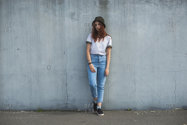 Full body of trendy teen red haired female student in casual outfit and hat leaning back on gray shabby concrete wall on urban street