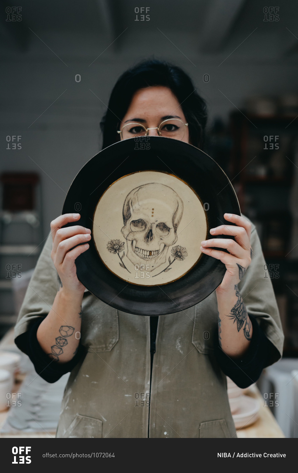 Creative female artisan in dirty clothes covering face with handmade clay plate with drawn skull and looking at camera