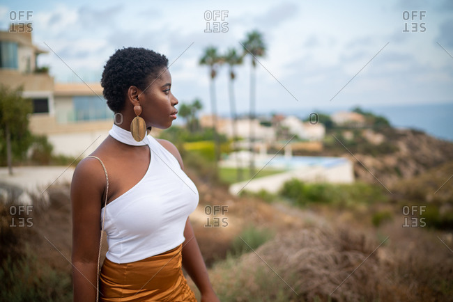 Trendy pondering African American female in earrings admiring ocean under cloudy sky while looking away