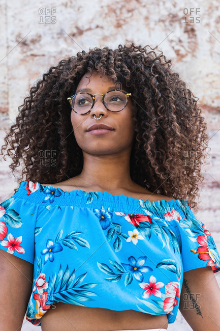 Stylish African American woman in glasses touching curly hair and looking away while standing near marble wall on street
