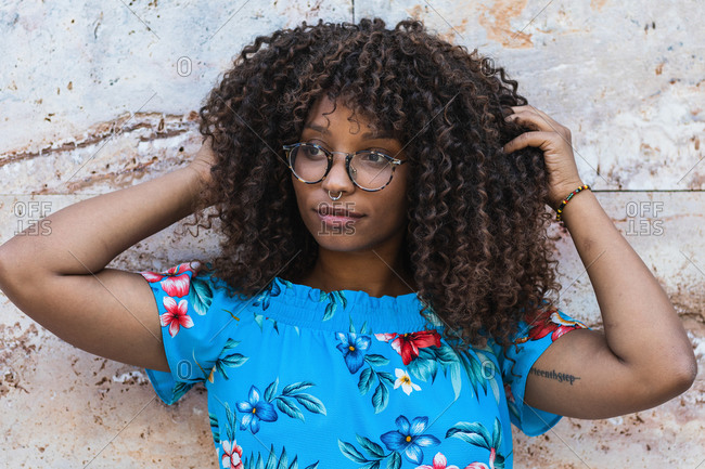Stylish African American woman in glasses touching curly hair and looking away while standing near marble wall on street