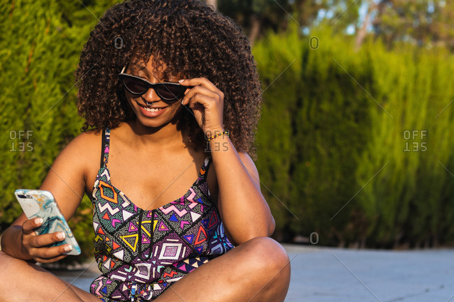 Young black woman in trendy outfit and sunglasses sitting cross legged on border and browsing smartphone on summer day