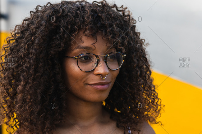Young black woman with glasses and curly hair while standing near vivid wall on city street