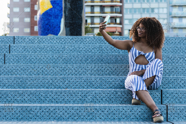 Full body positive African American female in striped apparel smiling and taking selfie while sitting on stairs on city street