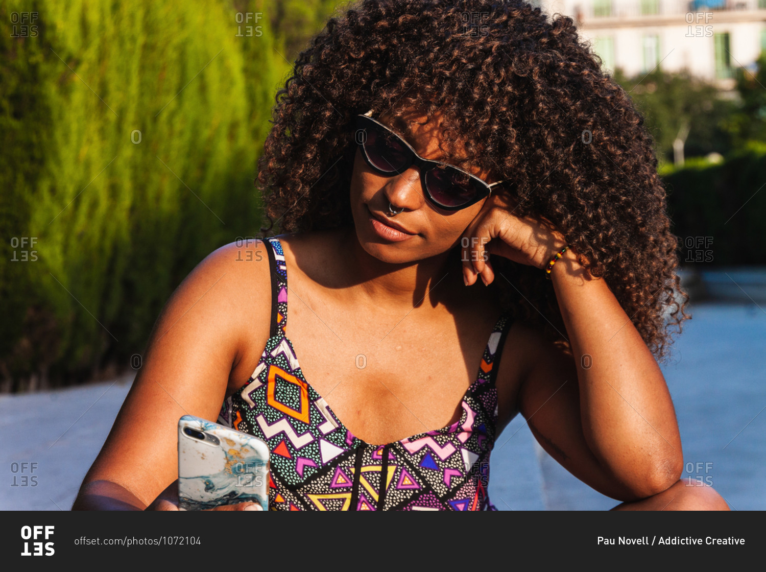 Young black woman in trendy outfit and sunglasses sitting cross legged on border and browsing smartphone on summer day