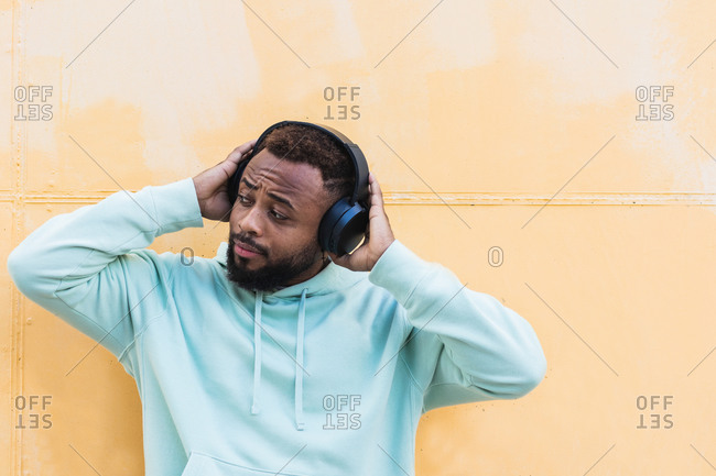 Adult bearded African American male in blue hoodie enjoying music with wireless headphones against yellow background