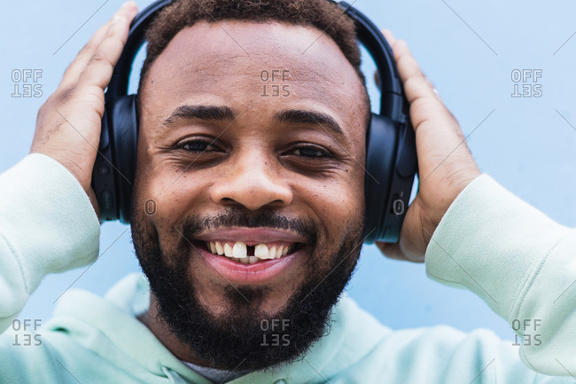 Happy adult bearded African American male in blue hoodie enjoying music with wireless headphones against blue background