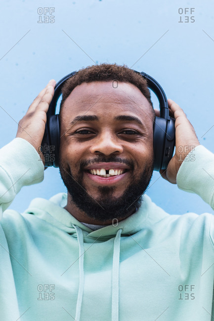 Happy adult bearded African American male in blue hoodie enjoying music with wireless headphones against blue background