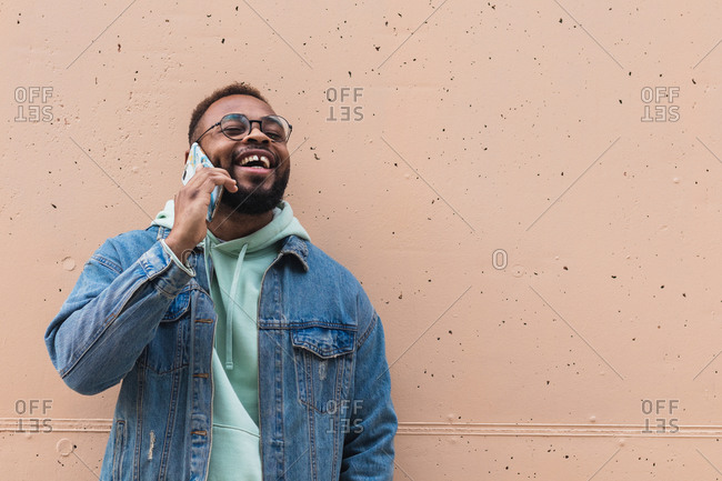 Adult hipster bearded African American male in casual outfit and glasses having phone conversation and laughing satisfied with news while standing against beige stone wall on street