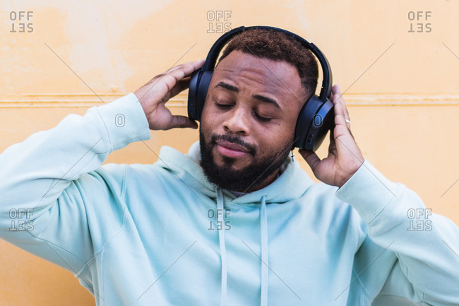 Happy adult bearded African American male in blue hoodie enjoying music with wireless headphones against yellow background