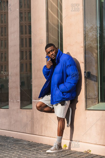 Confident African American male model wearing vivid blue jacket while standing on modern building on the street and looking away