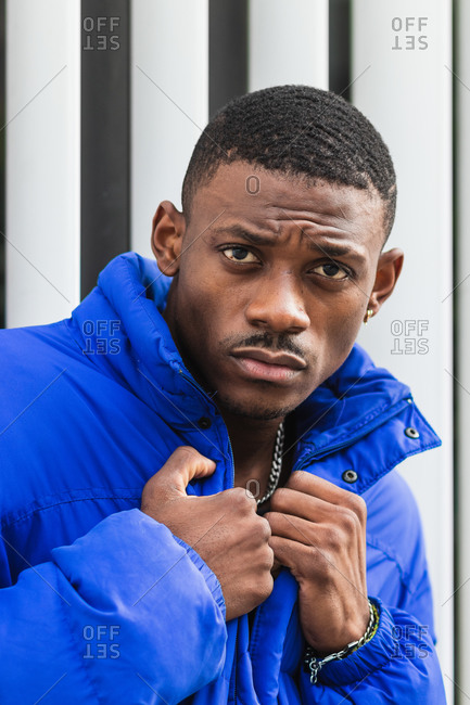 Side view of confident African American male model wearing vivid blue jacket while standing on metal wall on the street and looking at camera