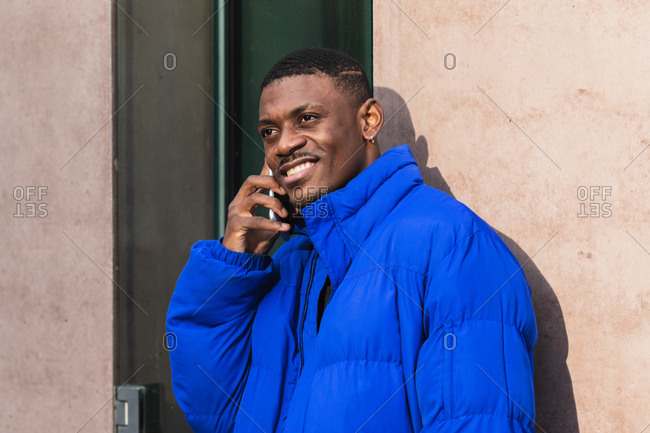 Positive African American male in warm jacket standing on street near building and messaging on social media via smartphone