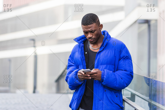 African American male in warm jacket standing on street near building and messaging on social media via smartphone
