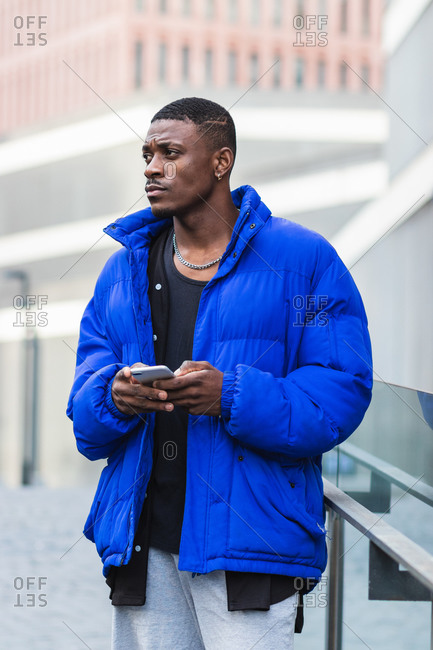 African American male in warm jacket standing on street near building and messaging on social media via smartphone looking away
