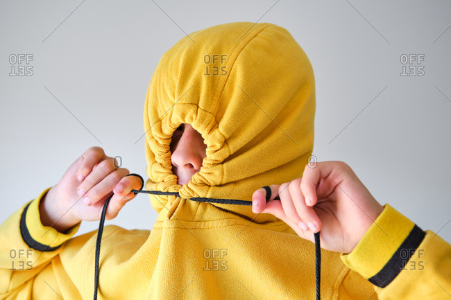 Anonymous expressive teenager hiding in yellow hoodie and looking at camera on gray background in studio