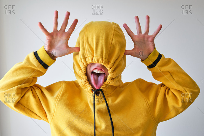Unrecognizable teen in vivid yellow hoodie covering face with hood and showing tongue while having fun on gray background in studio