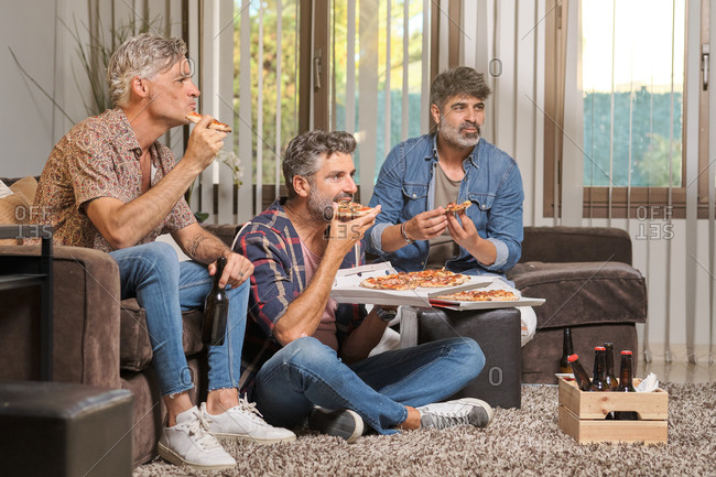 Full body of cheerful mature ethnic friends sharing pizza with beer bottles spending time together in cozy living room