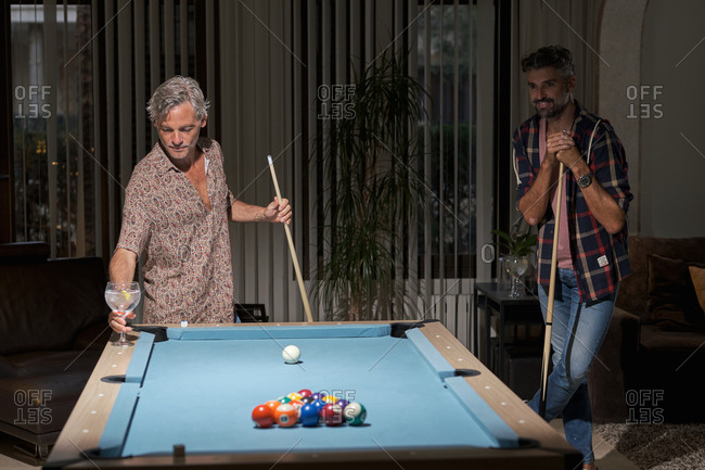 Happy mature friends in casual clothing playing pool and drinking cocktails while resting in cozy room in light of lamps