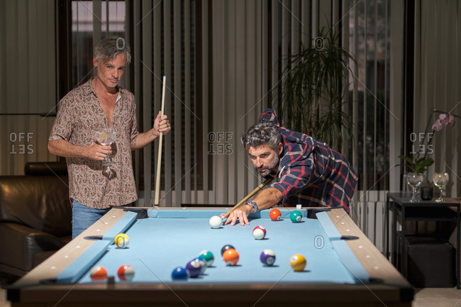 Happy mature friends in casual clothing playing pool and drinking cocktails while resting in cozy room in light of lamps