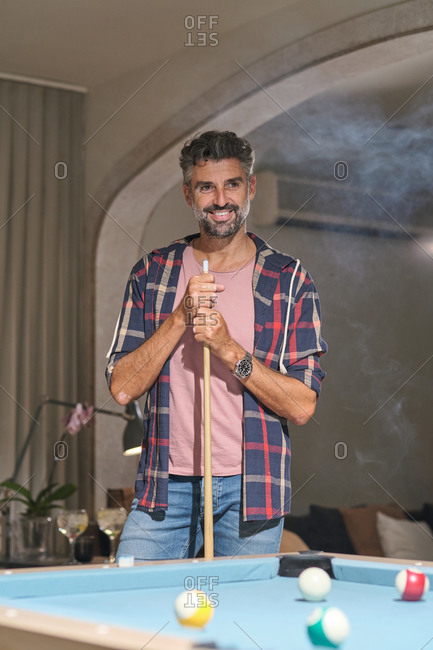 cheerful adult male with gray hair in casual clothing standing looking away with cue stick while playing billiard in bright room