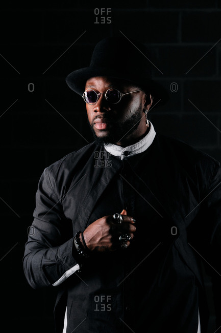 Determined African American male wearing round sunglasses and trendy black clothes standing on dark background in studio and looking away