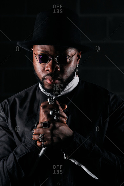 Determined African American male wearing round sunglasses and trendy black clothes standing on dark background in studio and looking at camera