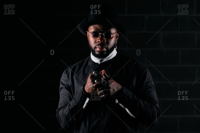 Determined African American male wearing round sunglasses and trendy black clothes standing on dark background in studio and looking at camera