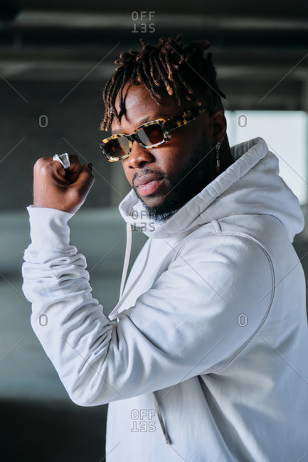 Side view of determined African American male in fancy outfit and sunglasses showing bicep and demonstrating power while seriously looking at camera
