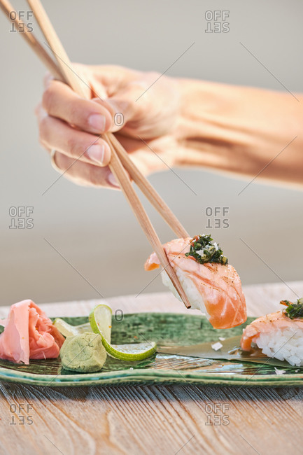 Cropped unrecognizable person hands holding chopstick eating delicious sushi Nigiri with salmon and greenery served on plate in Asian restaurant