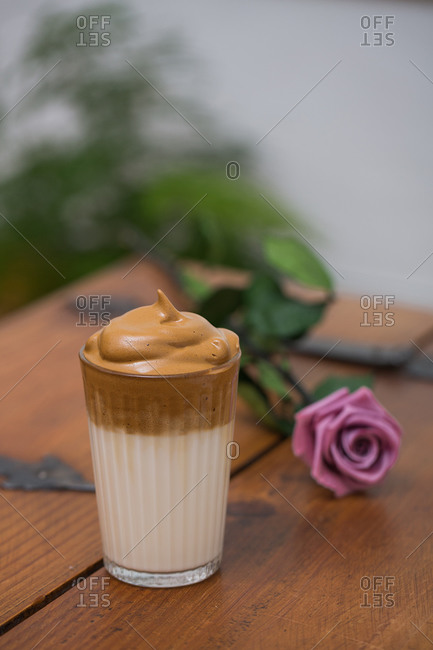 Glass of delicious cool Dalgona coffee placed on wooden table with rose flower in cafe