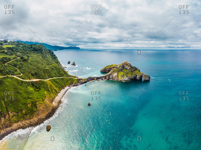Drone view of paving stone way leading along stone bridge and ridge of rocky hill to lonely house on island Gaztelugatxe surrounded by tranquil sea water under cloudy sky in Basque Country