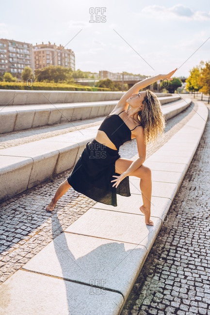 Full body of slim female in black sporty top and skirt standing on stone steps and doing Extended Side Angle Pose while practicing yoga on street